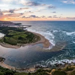Vue aérienne d'une rivière serpentant à travers des collines verdoyantes pour se jeter dans l'océan Atlantique sur une plage de sable au coucher du soleil.